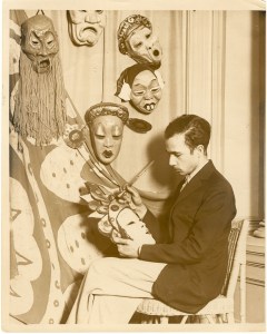 Ralph Chessé sits cross legged in a chair painting a mask with a crown on it.